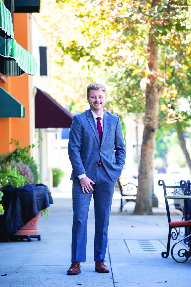 Sam Johnson of SMMA Insurance, smiling in a blue suit on a sidewalk beside café tables